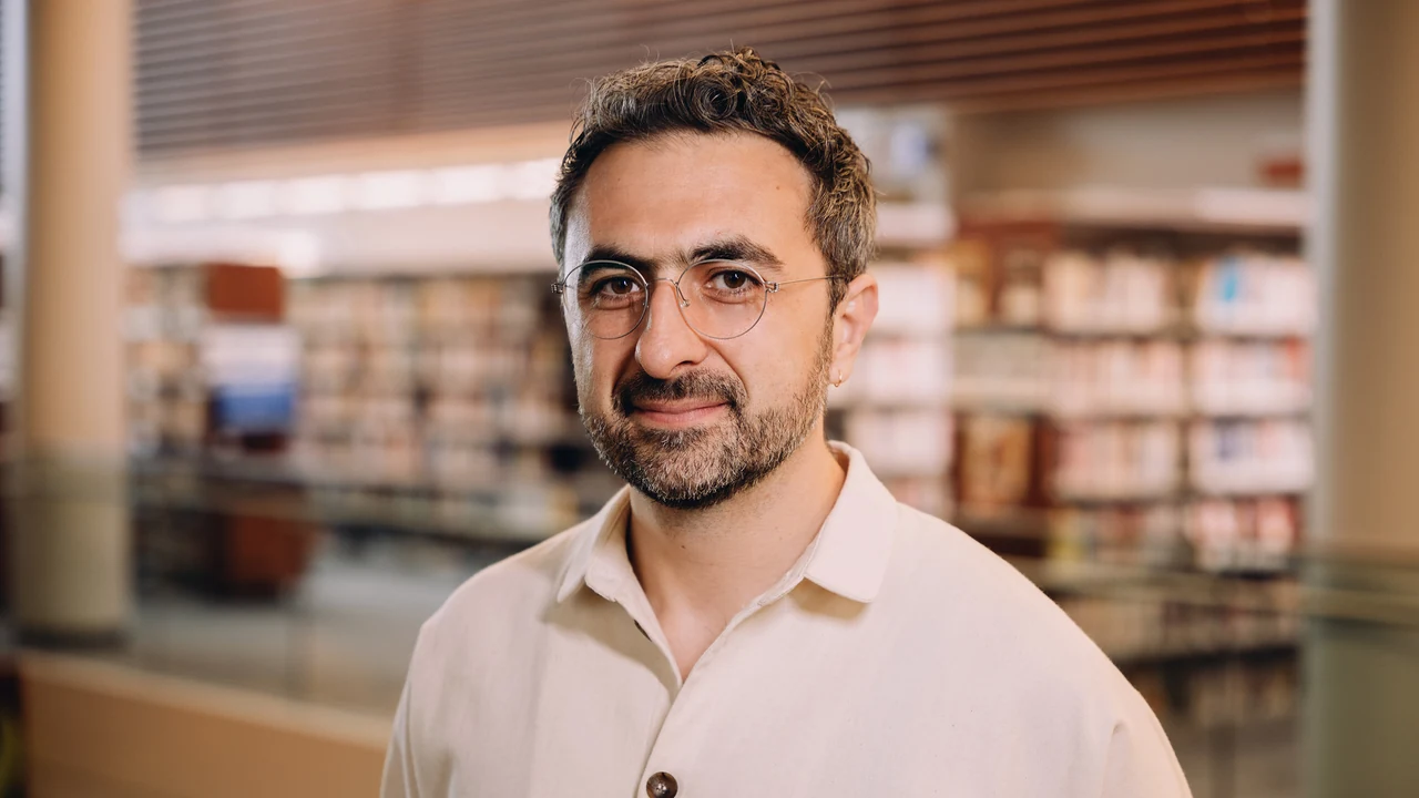 Microsoft AI CEO Mustafa Suleyman, in round glasses and a cream shirt against a blurred library bookshelf background
